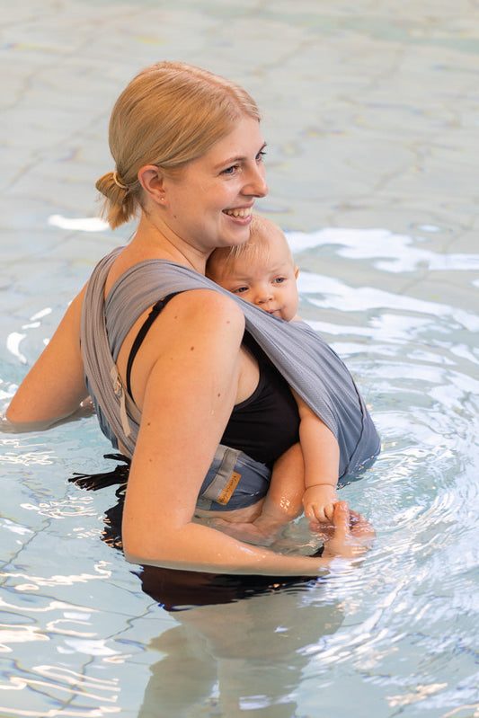 Frau trägt Baby in AQUA Loops in "ocean", sie steht seitlich bis zur Hüfte im Wasser.