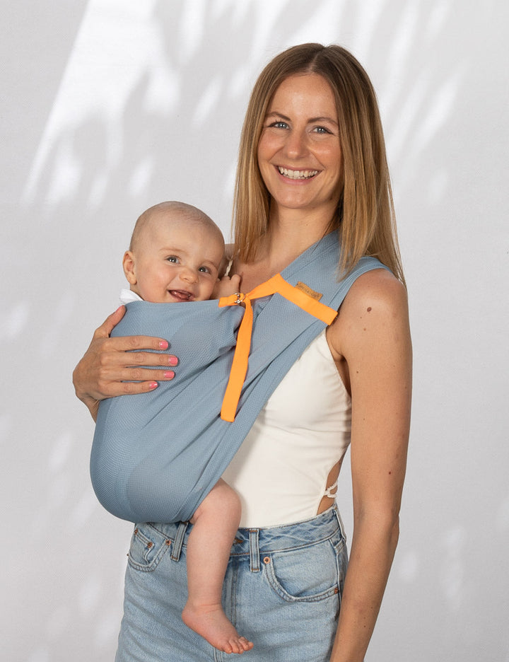 Frau trägt Baby mit einem AQUA Loop in "ocean mit orangenem Ripsband" als Hüftsitz auf der Hüfte. Beide schauen in die Kamera und sie hält eine Hand am Rücken des Babys. 