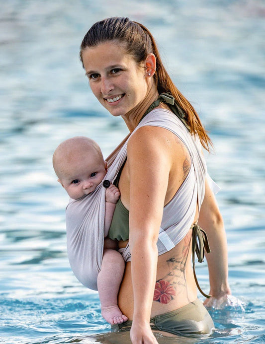 Frau trägt Baby in AQUA Loops in "sand", sie steht seitlich bis zur Hüfte im Wasser und schaut in die Kamera.