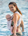Frau trägt Baby in AQUA Loops in "sand", sie steht seitlich bis zur Hüfte im Wasser und schaut in die Kamera.